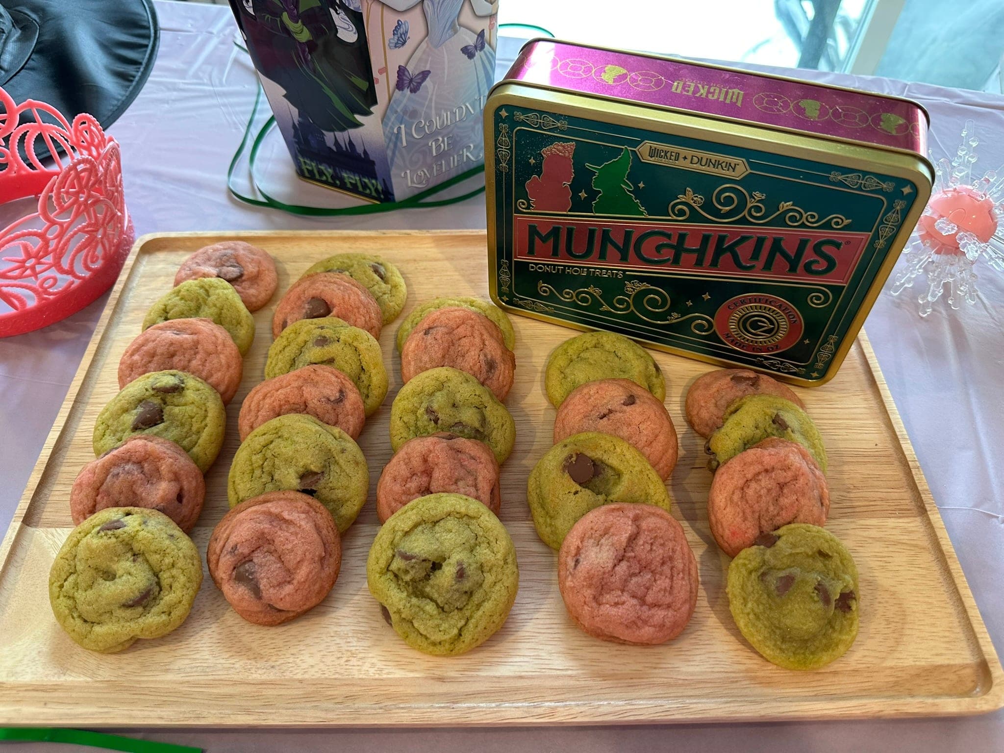 Two dozen chocolate chunk cookies in pink and green, laid out in rows on a wooden board on a Wicked-themed party table